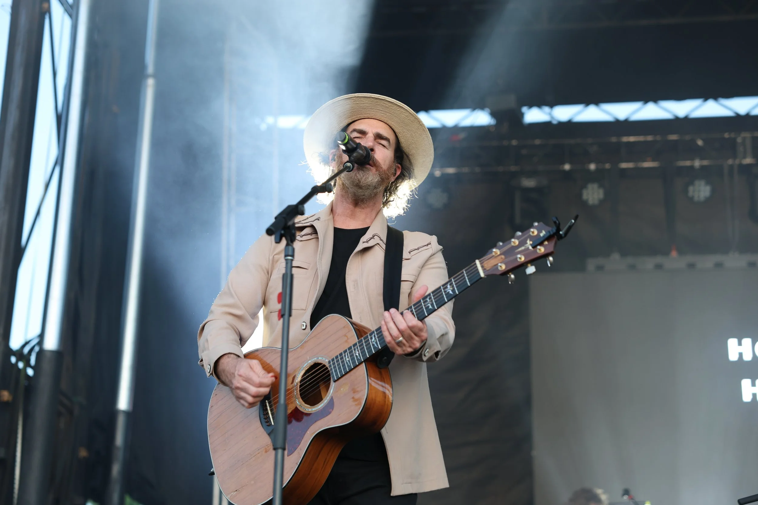 A man with a beard, wearing a beige jacket, black shirt, and a wide-brimmed hat, singing into a microphone and playing an acoustic guitar on a stage.