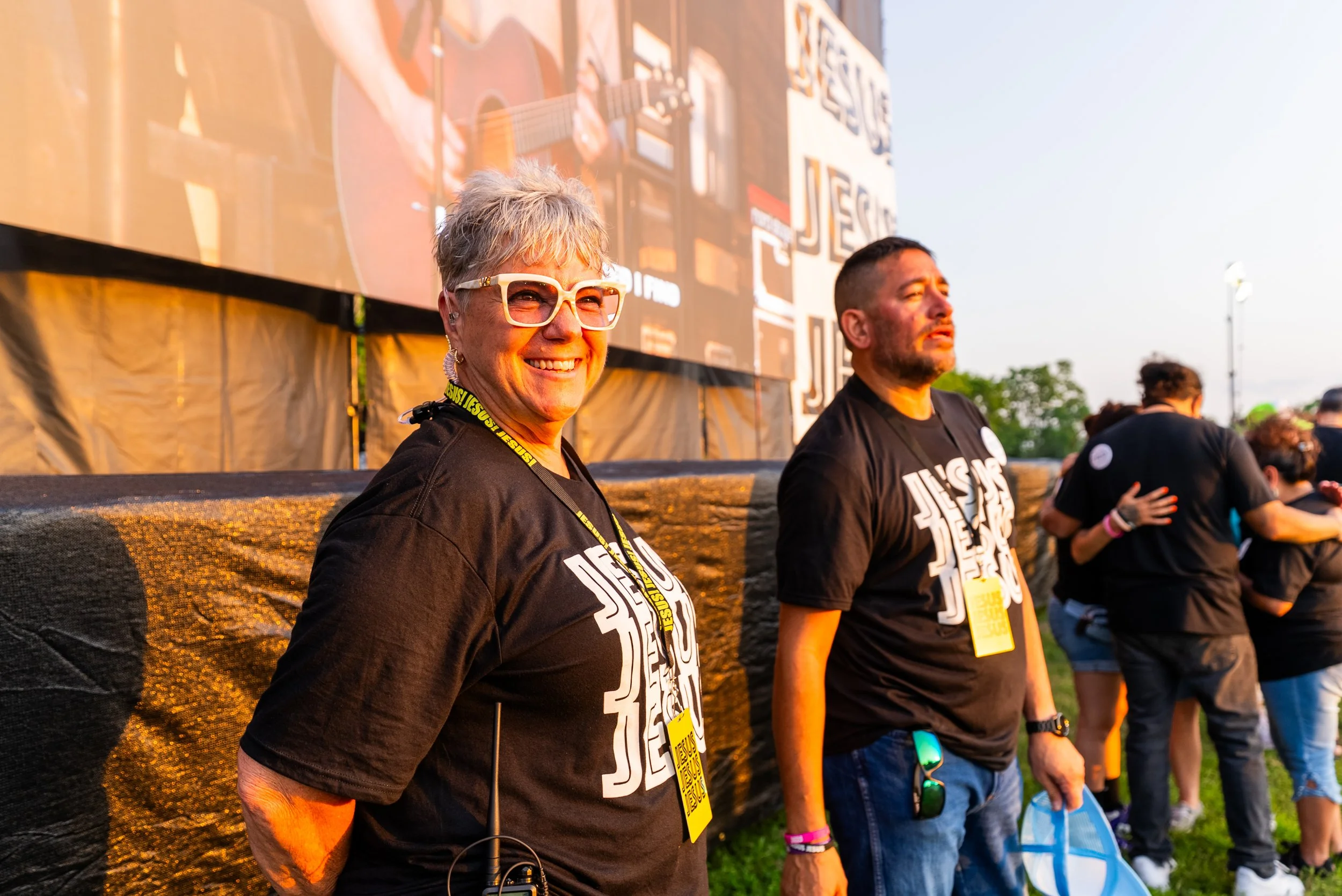Smiling woman with short gray hair, glasses, and a black shirt, standing outdoors during sunset at a music or event festival, with a man in a black shirt and others in the background.