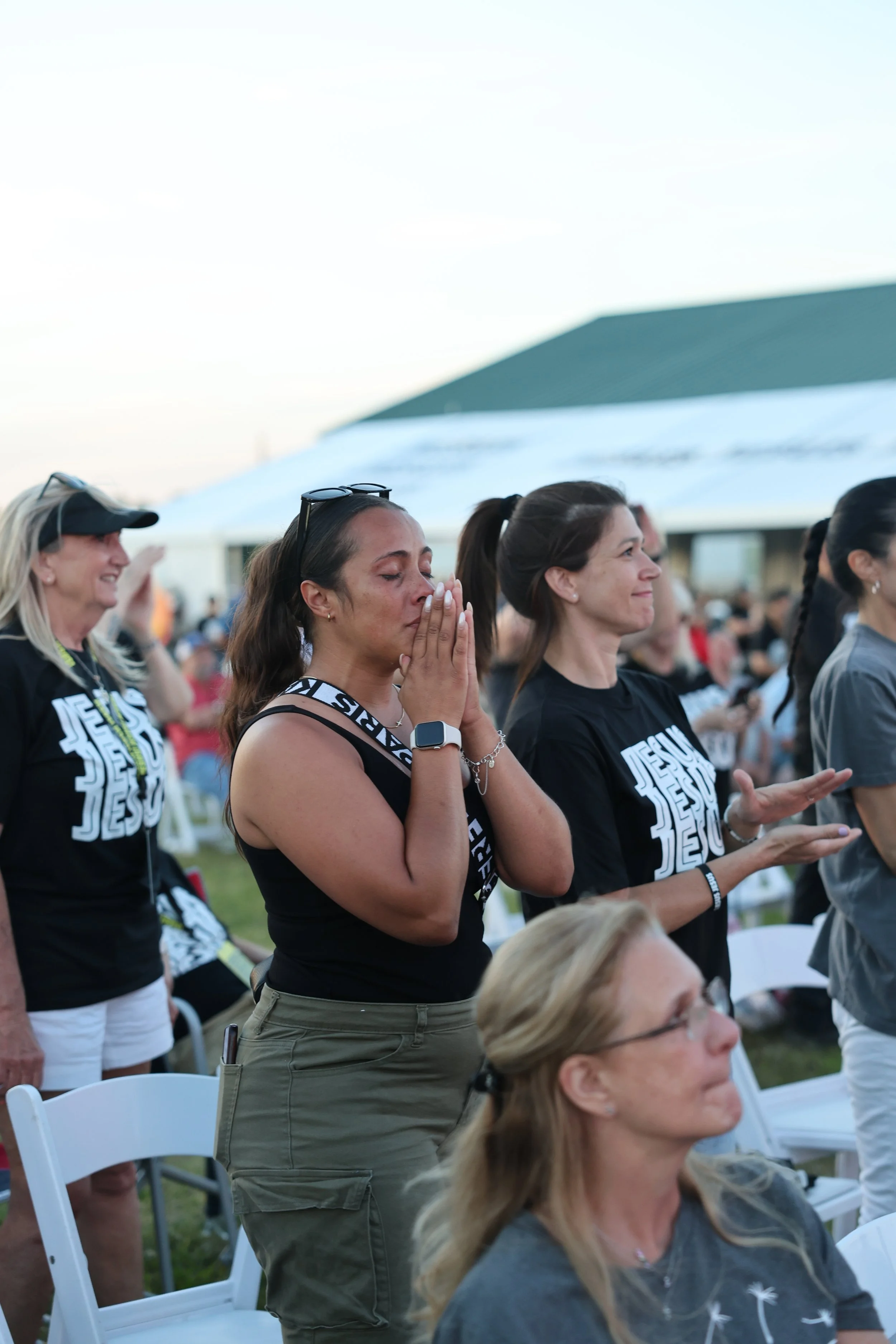 A group of women attending an outdoor religious event, some with eyes closed in prayer or reflection, others smiling, with chairs and tents in the background.