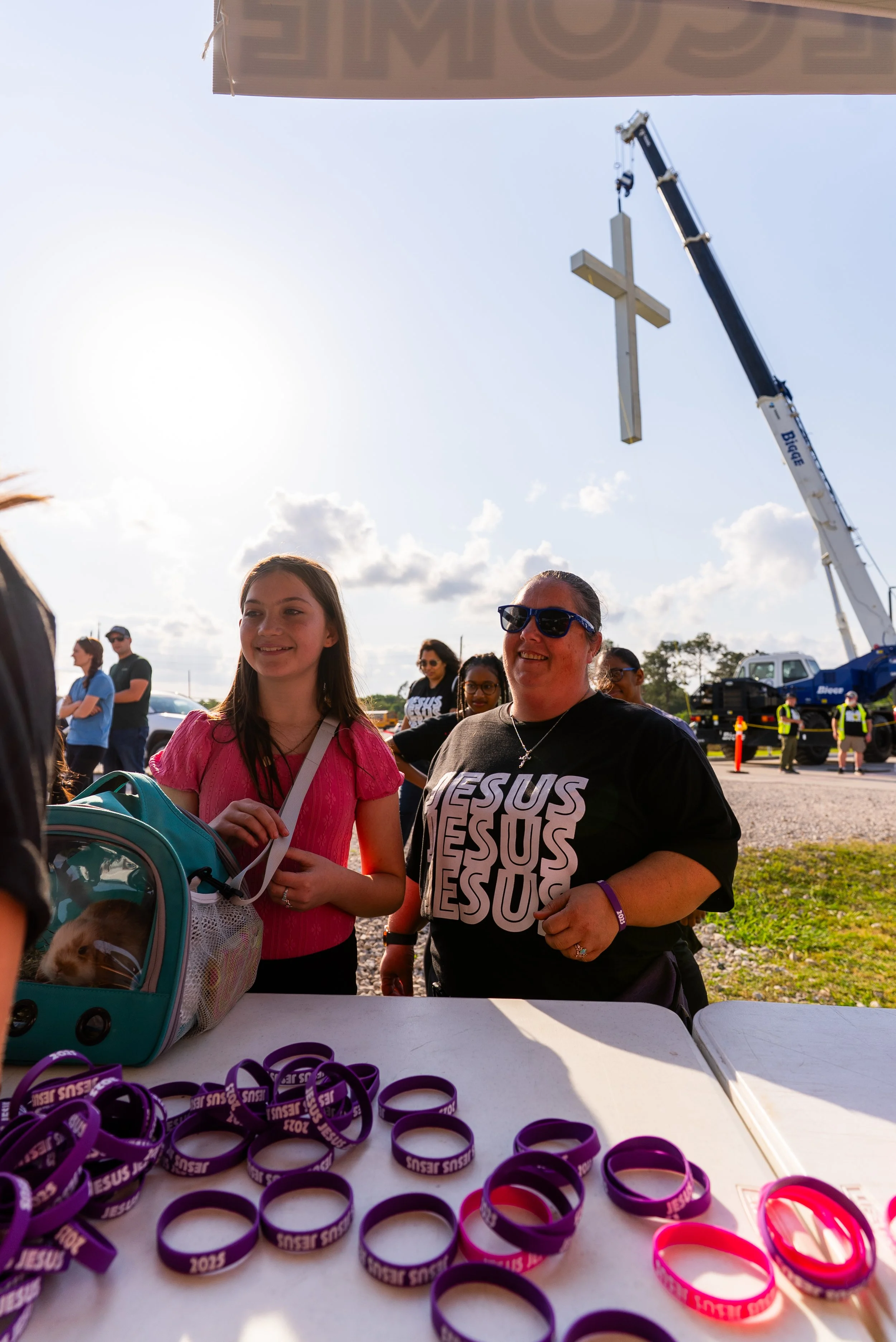 Women and children at a religious event table, with a large cross hanging from a crane in the background and purple wristbands on the table.