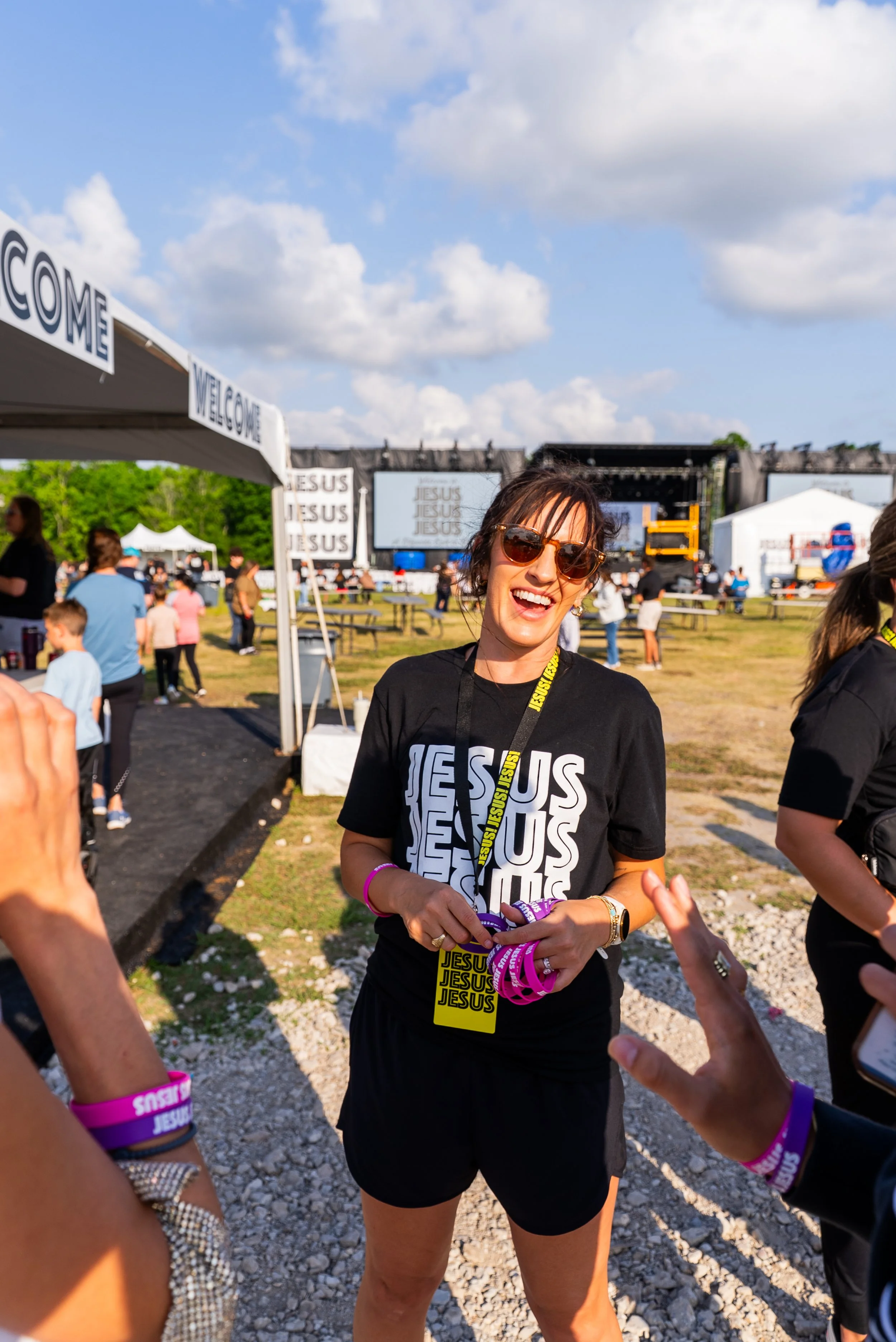 Young woman wearing sunglasses and a black T-shirt with "JESUS" printed repeatedly, standing outdoors at a large religious event with a stage and crowd in the background, smiling and holding purple wristbands.