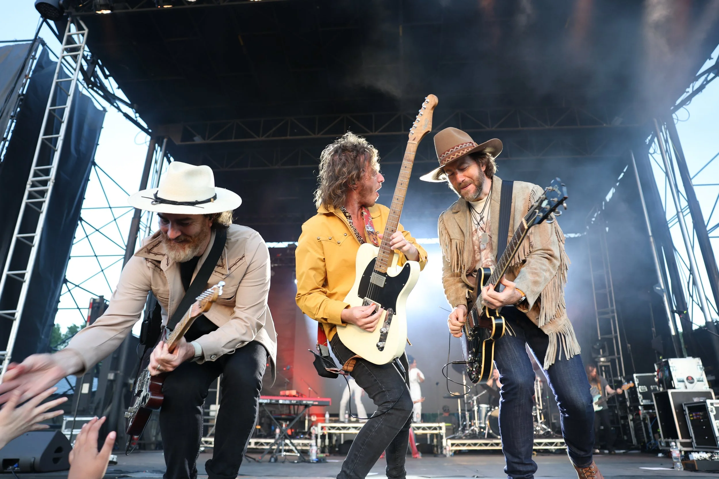 Three musicians playing guitars on stage during a concert, with audience hands reaching up in front of them.
