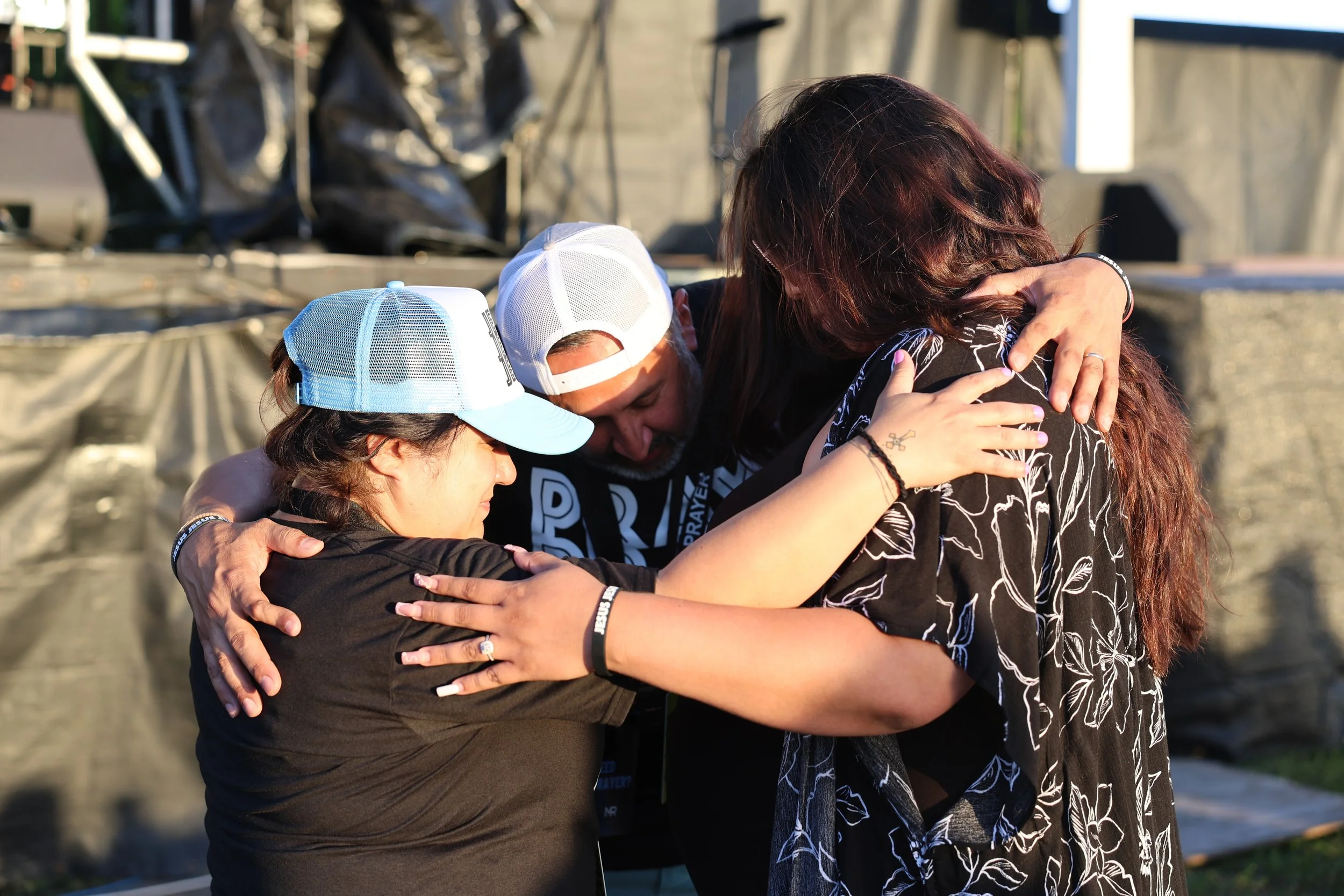 Three people embracing each other in a group hug outdoors, with two women wearing caps and dressed casually, and a man with a beard, all showing a moment of connection and support.