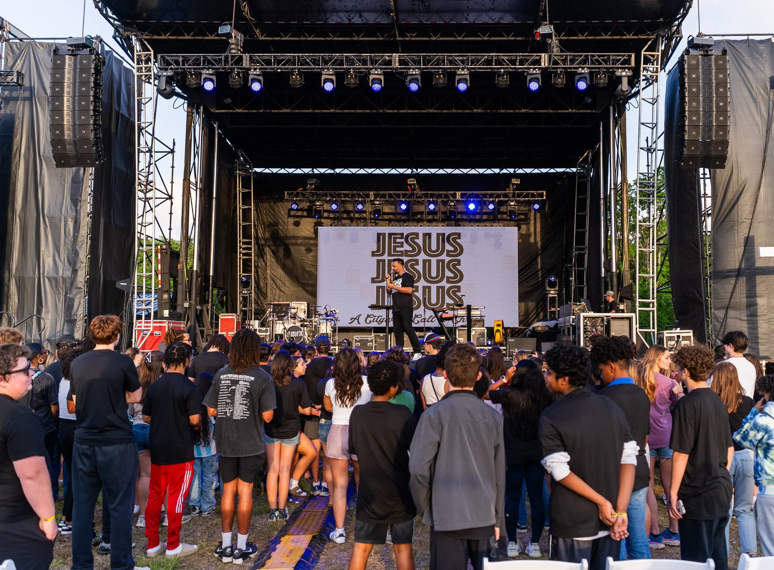 A large outdoor concert stage with a crowd of young people watching a performance. The stage displays a screen with the words 'JESUS' repeated three times, followed by 'A City Called Heaven'. The performers are on stage with musical instruments and s