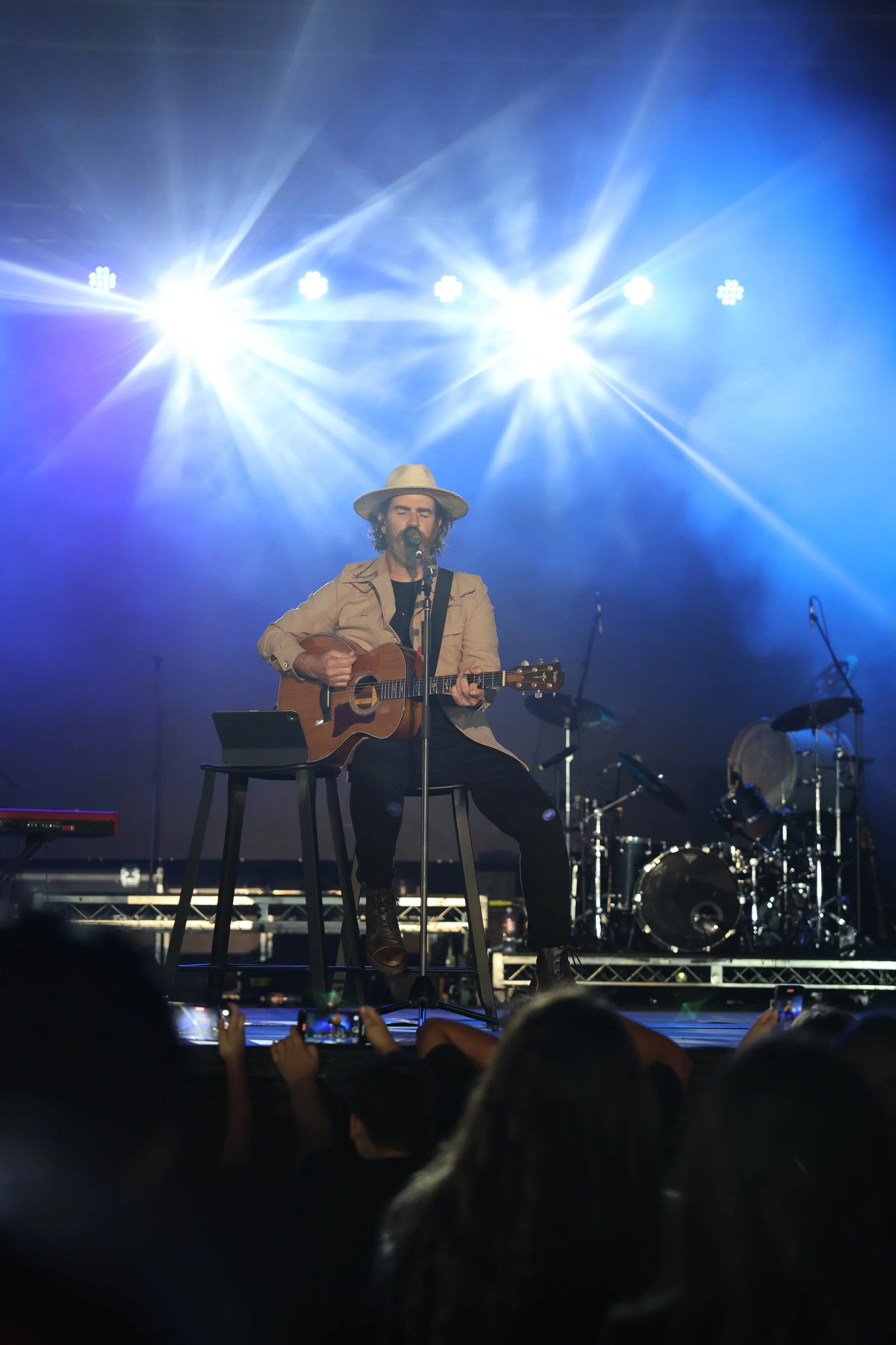 A man with a beard and long hair, wearing a wide-brimmed hat and beige jacket, is sitting on a stool on a stage, playing an acoustic guitar and singing into a microphone. There are bright stage lights shining behind him, with a drum set and other mus
