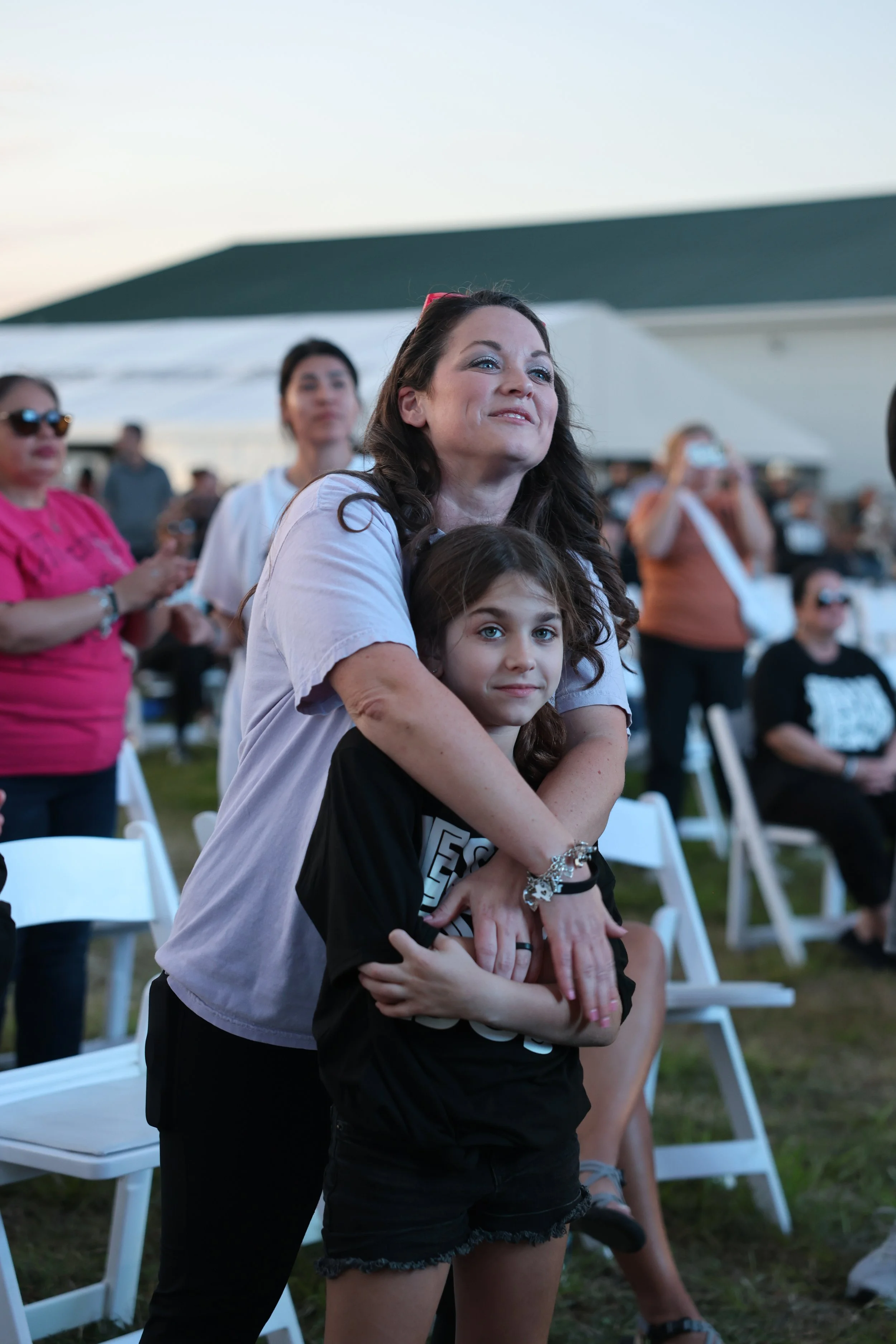 A woman and a young girl hugging at an outdoor event with many people seated and standing in the background.