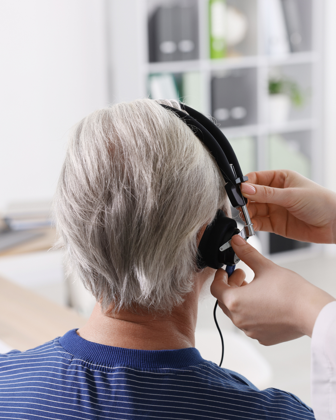 An elderly person with gray hair wearing a striped blue shirt listening with headphones in an indoor setting.