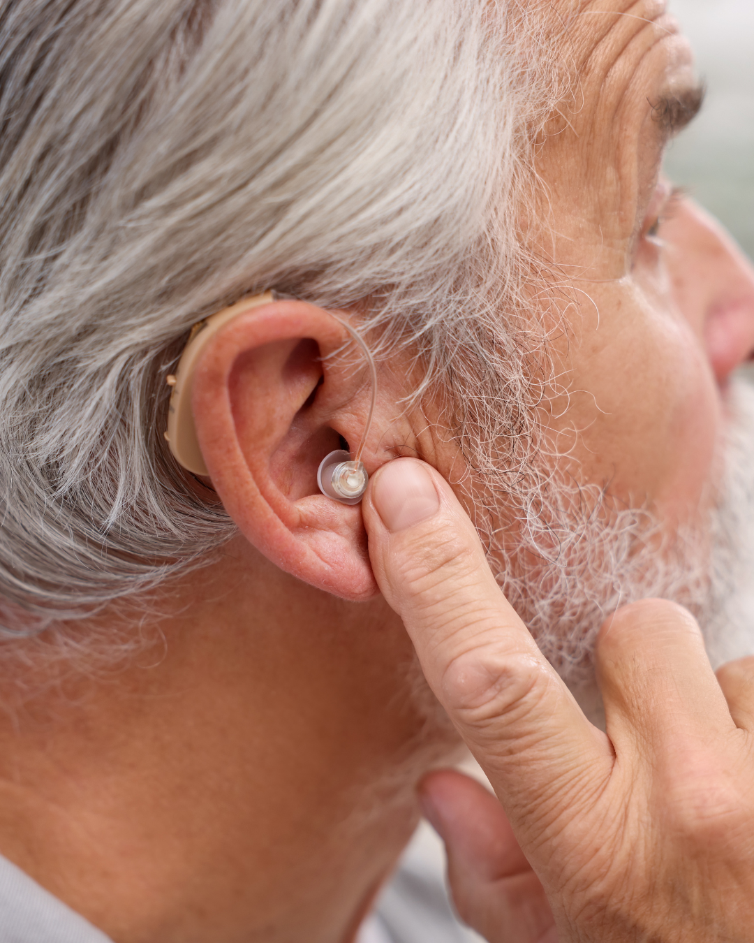 A close-up of an elderly man with gray hair inserting a hearing aid into his ear.
