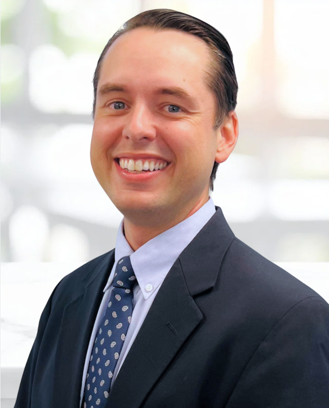 A smiling man in a dark suit, light blue shirt, and patterned tie standing in a bright, modern office environment.