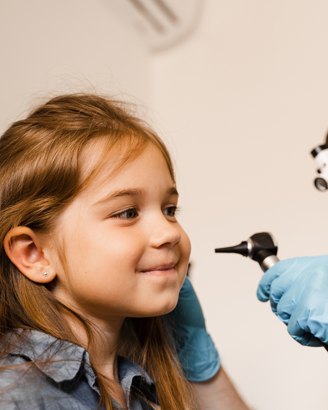 A young girl with light brown hair, wearing a blue shirt and earrings, smiling slightly as she looks into a medical device held by a person wearing blue gloves.