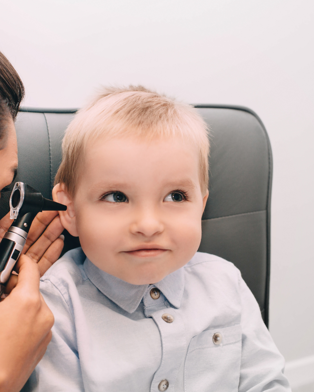 A young boy with light hair and blue eyes getting his ear examined with an otoscope at a doctor's office.