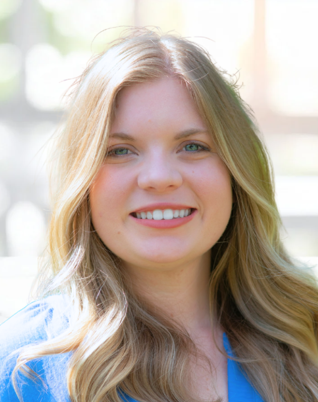 Close-up of a young woman with long, blonde hair and blue eyes smiling indoors with sunlight in the background.