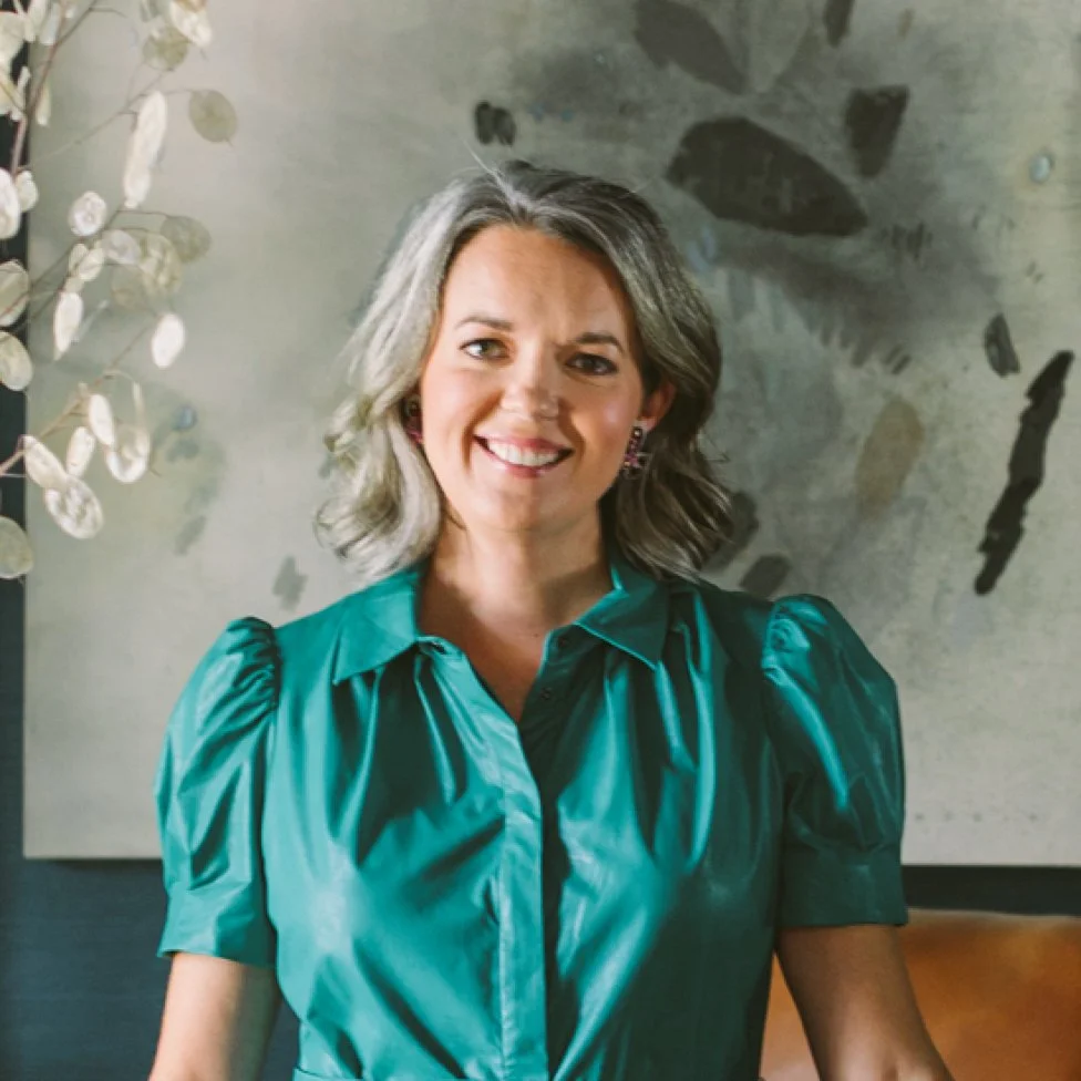 A woman with gray hair smiling, wearing a teal blouse with puffed sleeves, sitting in front of a textured wall and a piece of abstract art.
