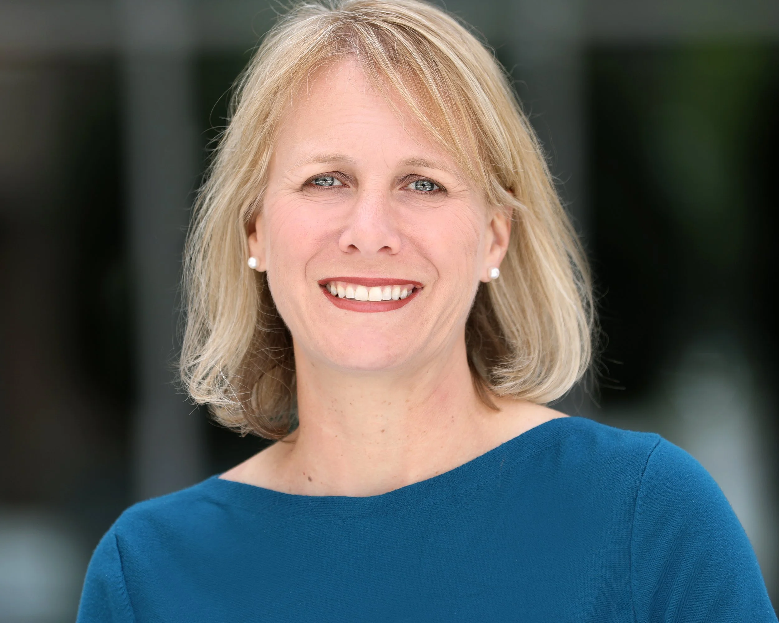 A woman with shoulder-length blonde hair, wearing pearl earrings and a blue top, smiling outdoors with a blurred background.