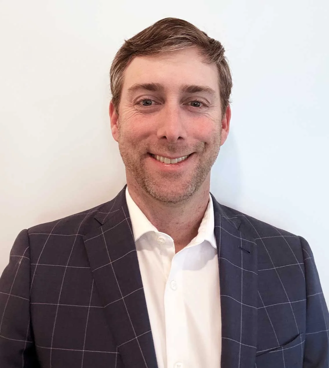 A smiling man in a checkered dark blazer and white shirt standing against a plain white wall.