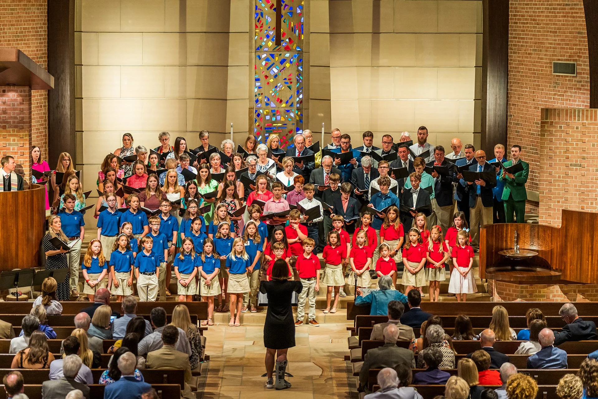 A large choir of children and adults performing in a church or auditorium, directed by a woman in black standing in front of them, with an audience seated in pews watching.