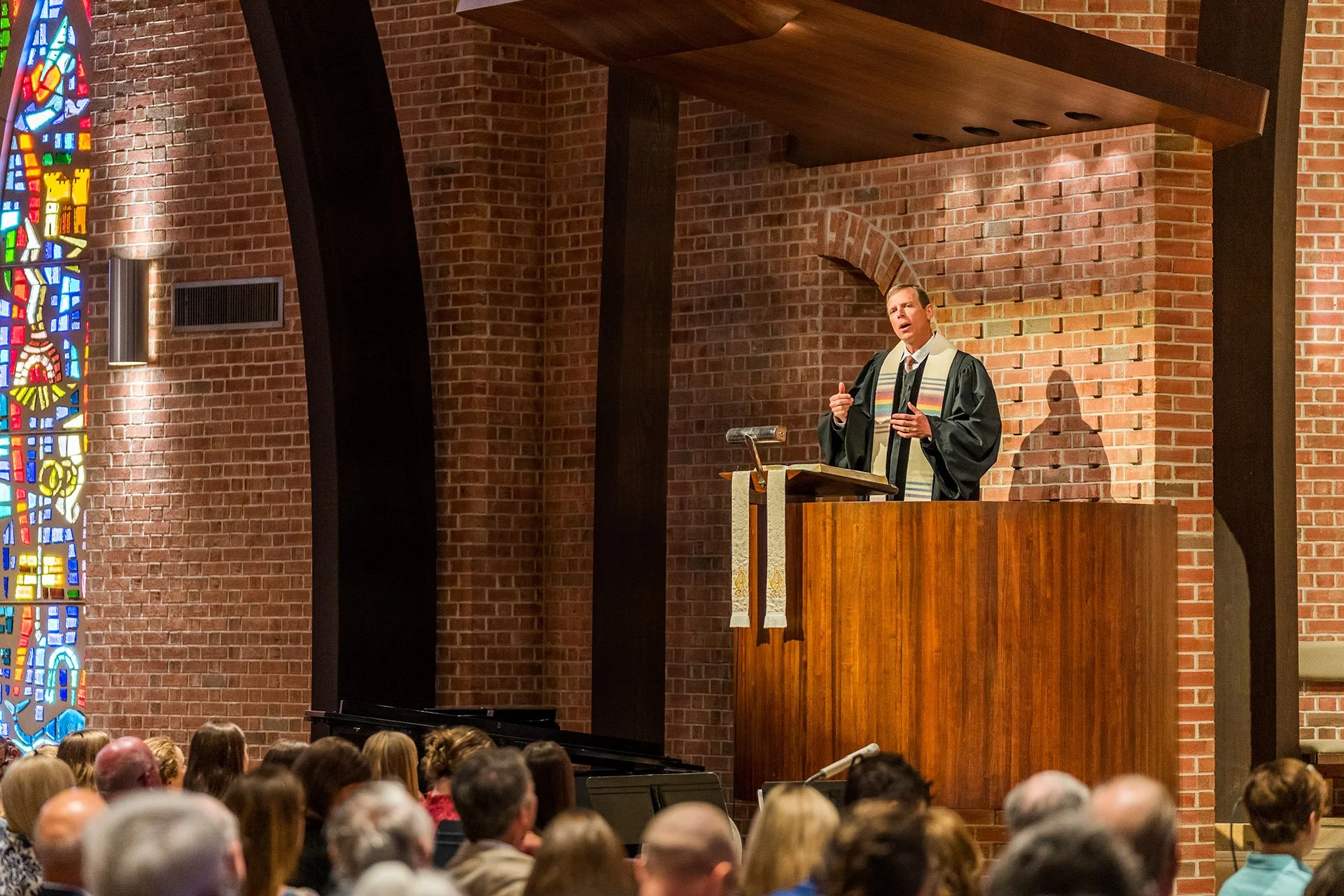 Pastor Ben Dorr is speaking in a pulpit at Westminster Presbyterian Church with brick walls and stained glass windows, addressing a congregation of seated people.