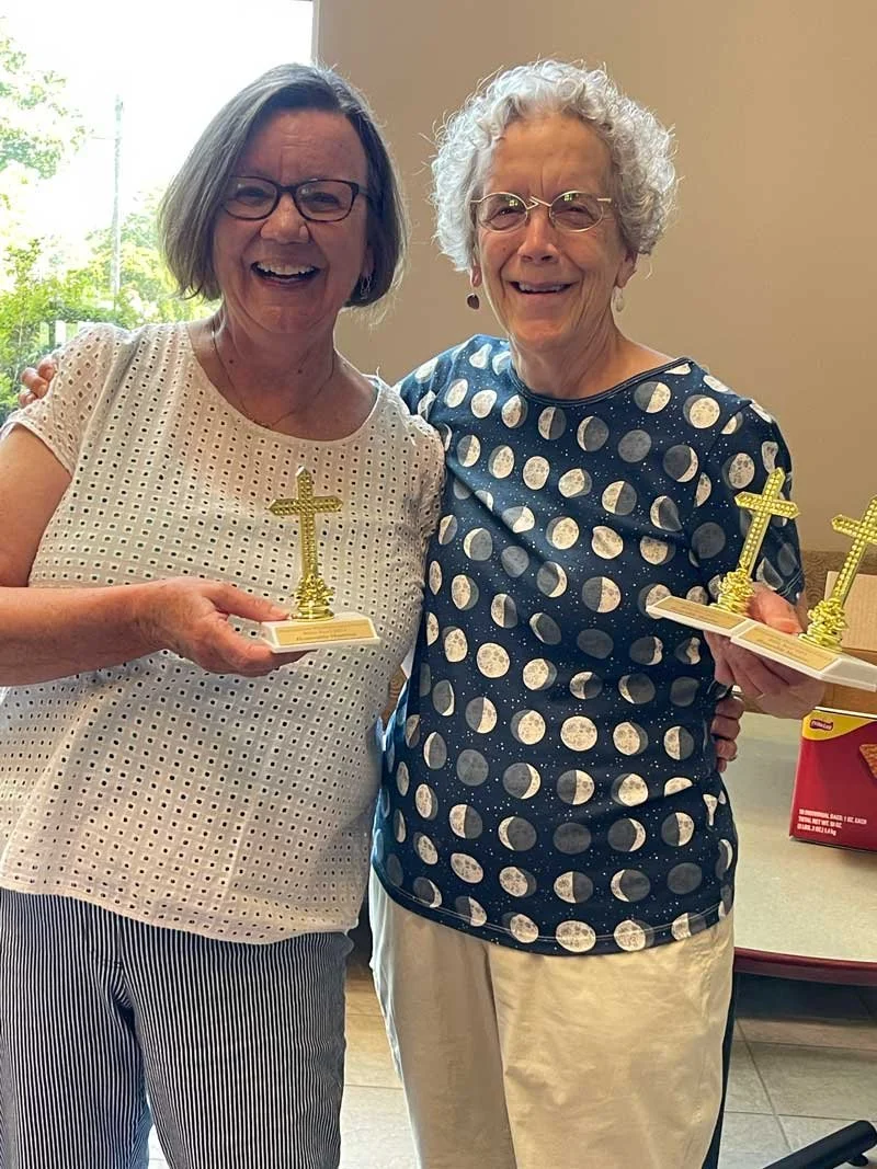 Two smiling women holding small gold cross trophies, standing indoors near a window with trees outside.