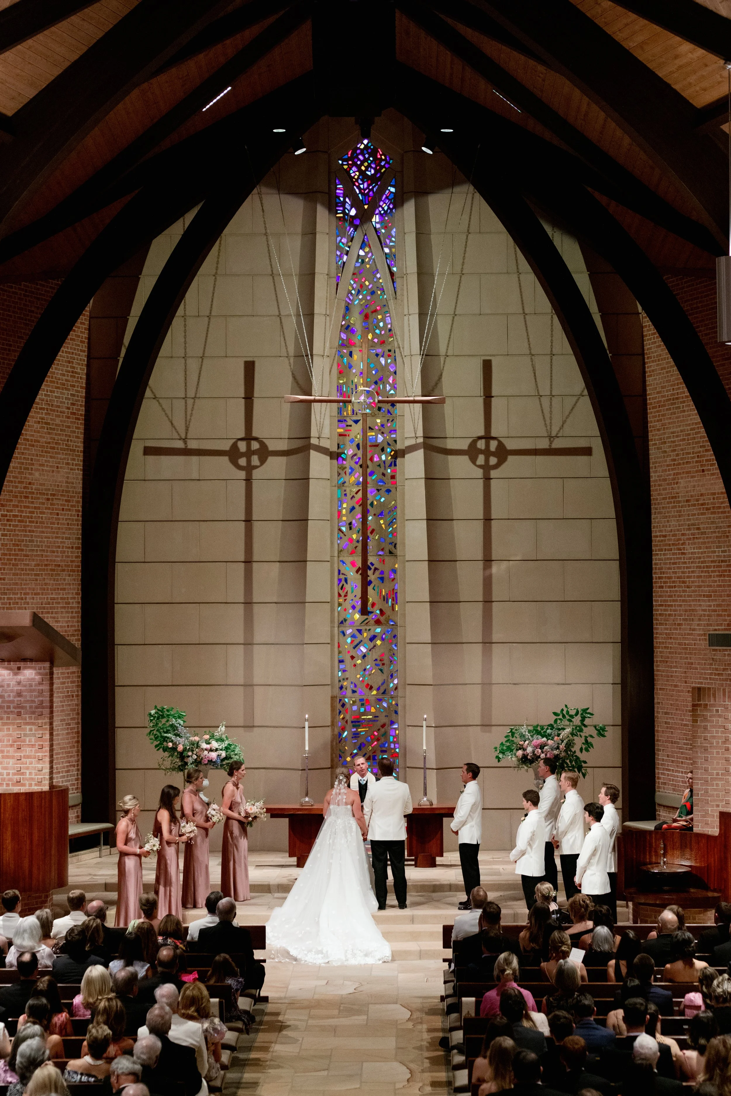 A wedding ceremony inside a church with a bride and groom exchanging vows at the altar, accompanied by bridesmaids and groomsmen. The church features a large stained glass window and a wooden ceiling.