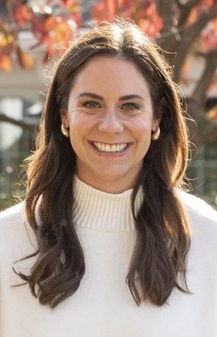 A woman with long brown hair, smiling, wearing a white sweater, outdoors with autumn-colored leaves in the background.