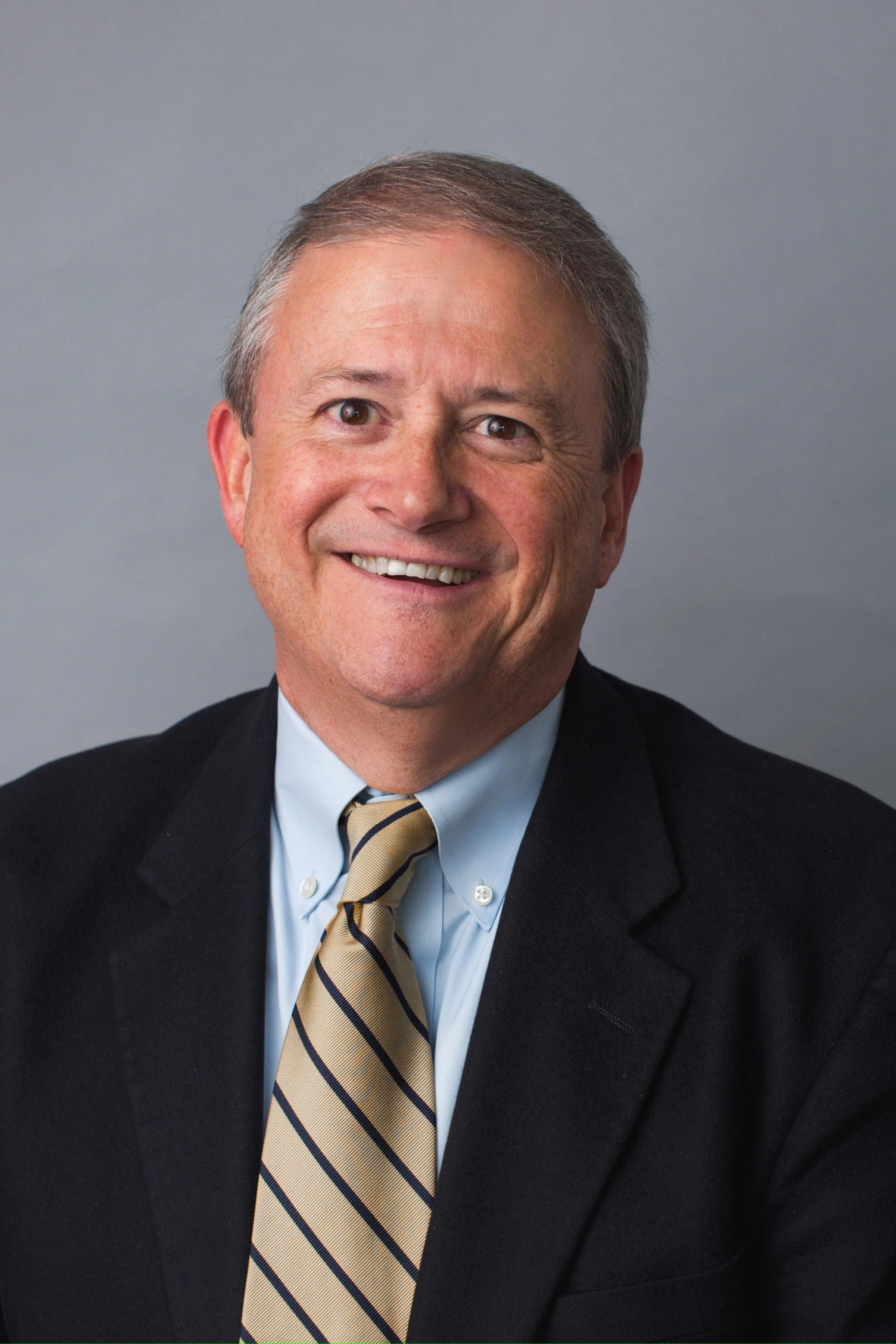 A professional headshot of a middle-aged man with short gray hair, wearing a dark suit, light blue shirt, and a beige striped tie, smiling against a plain gray background.