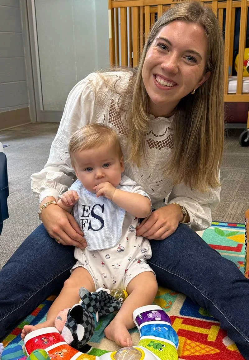 A woman with long blonde hair smiling and sitting on a colorful mat, holding a baby with light hair and blue eyes, who is sitting on her lap and sucking on their fingers, in a room with wooden furniture.