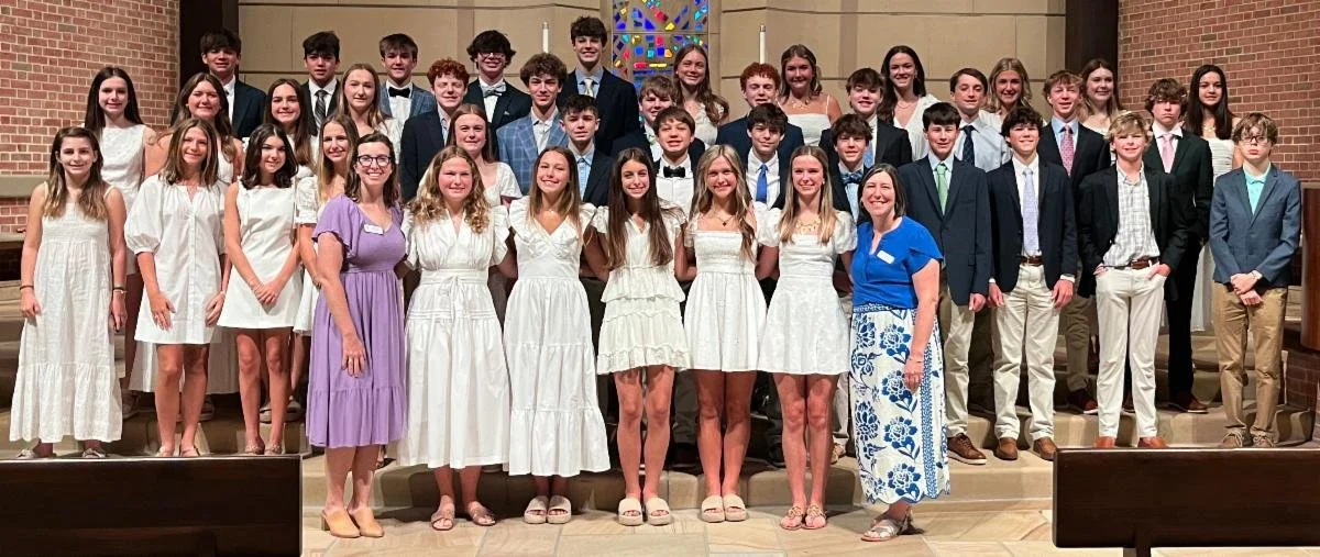 A large group of teenagers and two adult women posing for a photo inside a church or chapel. The teenagers are dressed in semi-formal attire with the boys in suits and the girls in white dresses. The woman on the right is wearing a blue top and a long skirt with blue and white patterns, while the woman on the left is also wearing a purple dress. The background features brick walls and stained glass windows.