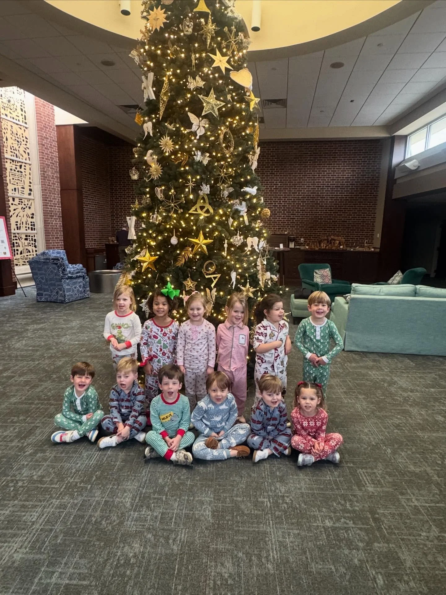 Group of young children sitting and standing in front of a decorated Christmas tree with lights and ornaments inside a lobby or common area.