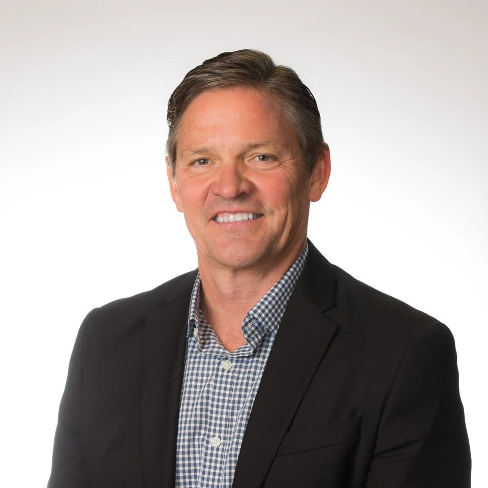 Headshot of a middle-aged man with brown hair, wearing a dark suit jacket and a checkered shirt, smiling against a plain white background.