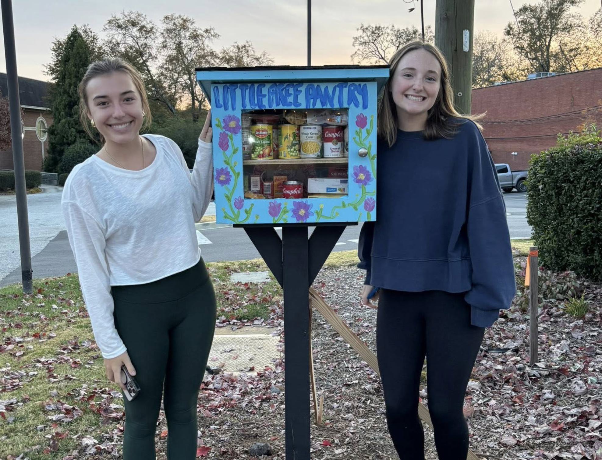 Two young women standing outside near a Little Free Pantry decorated with blue paint and purple flowers, smiling at the camera, during early evening.