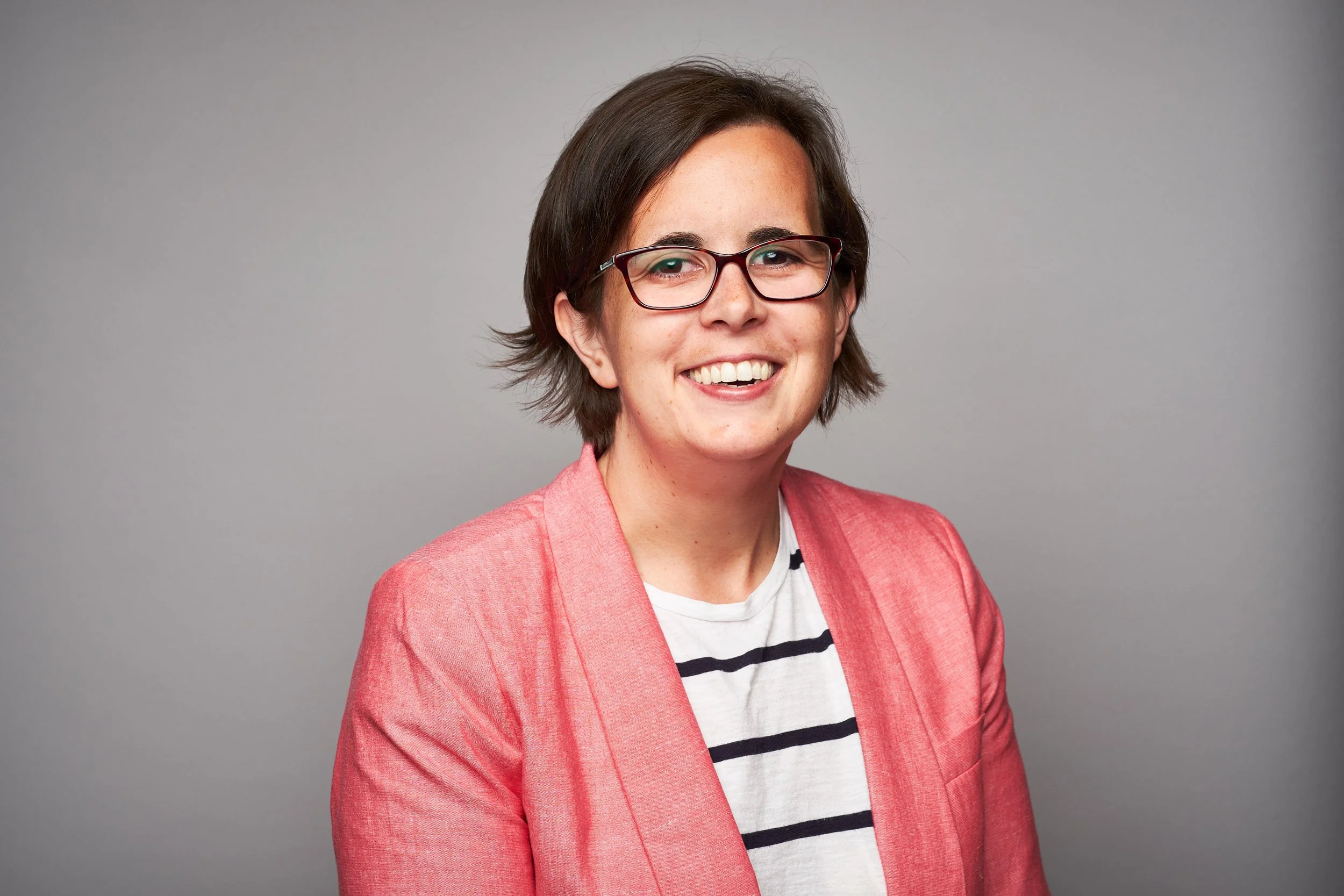 A woman with short brown hair, glasses, and a bright smile, wearing a pink blazer over a white and black striped shirt, standing against a plain gray background.