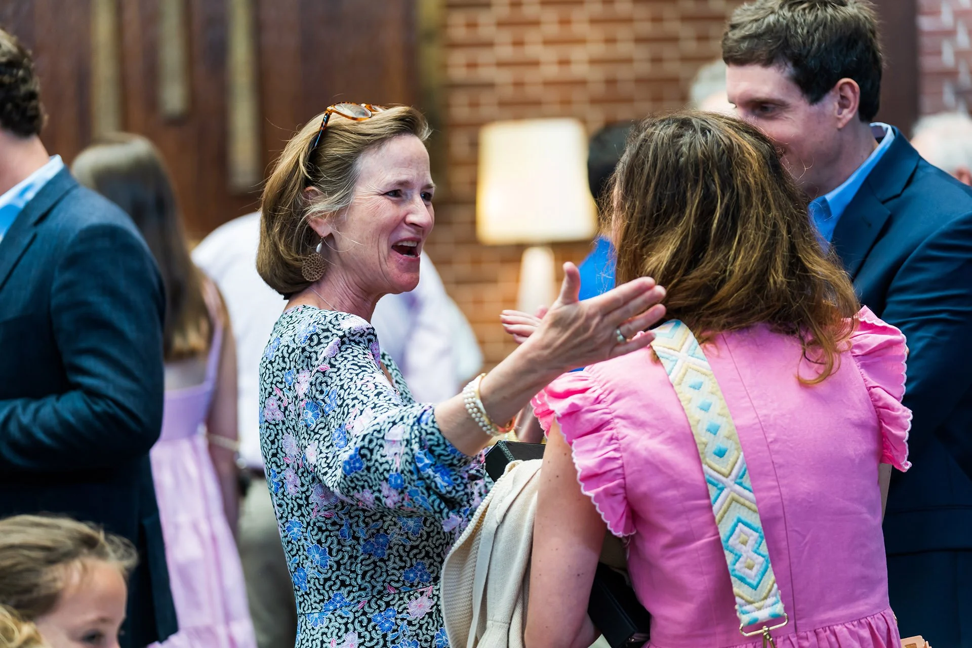 Three people engaged in conversation at an indoor social event, with a woman in a floral top speaking animatedly to a woman in a pink dress and a man in a blue jacket listening.