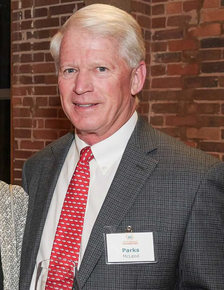 A middle-aged man with blond hair and blue eyes, dressed in a white shirt, red patterned tie, and gray checkered blazer, standing indoors against a brick wall. He is wearing a name badge that reads 'Parks McLeod'.