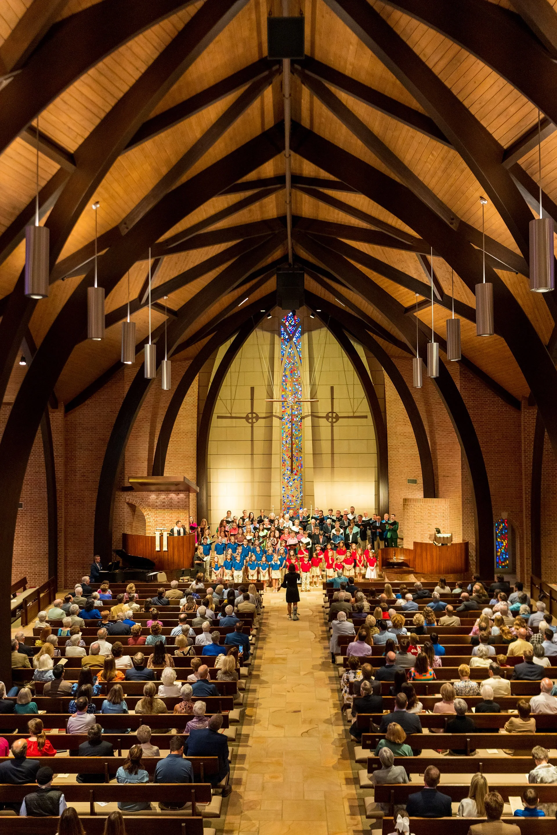 A choir singing in a church with an audience seated in pews. The church has wooden vaulted ceilings, hanging cylindrical lights, stained glass windows, and brick walls.