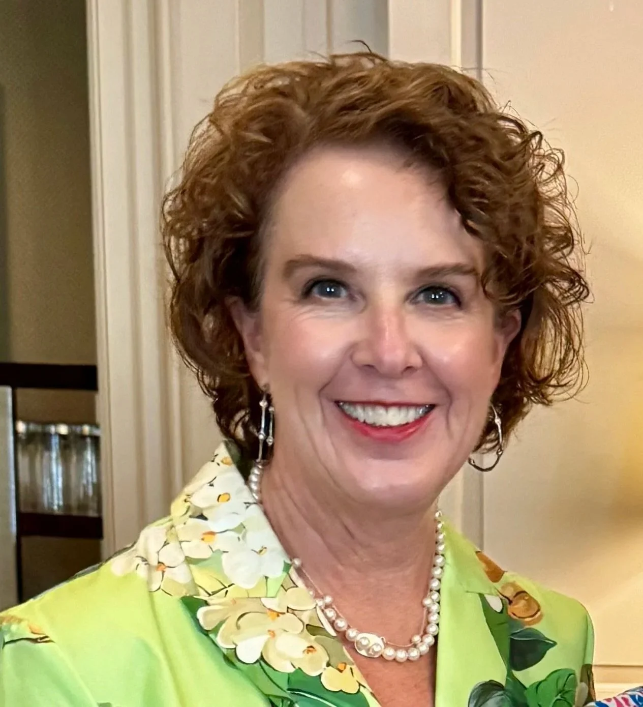 A smiling woman with short, curly red hair wearing a green floral blouse, pearl necklace, and earrings, standing indoors near a light-colored wall.