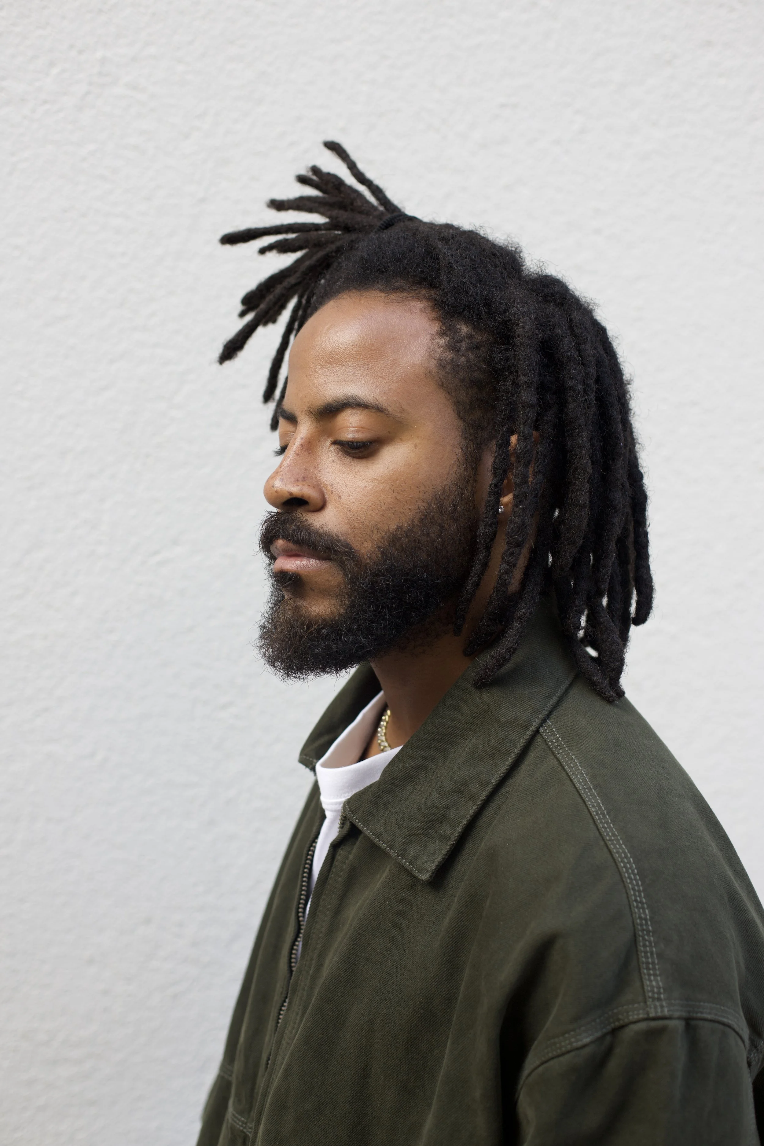 A man with dreadlocks and a beard looking down, wearing a dark green jacket against a plain white wall.