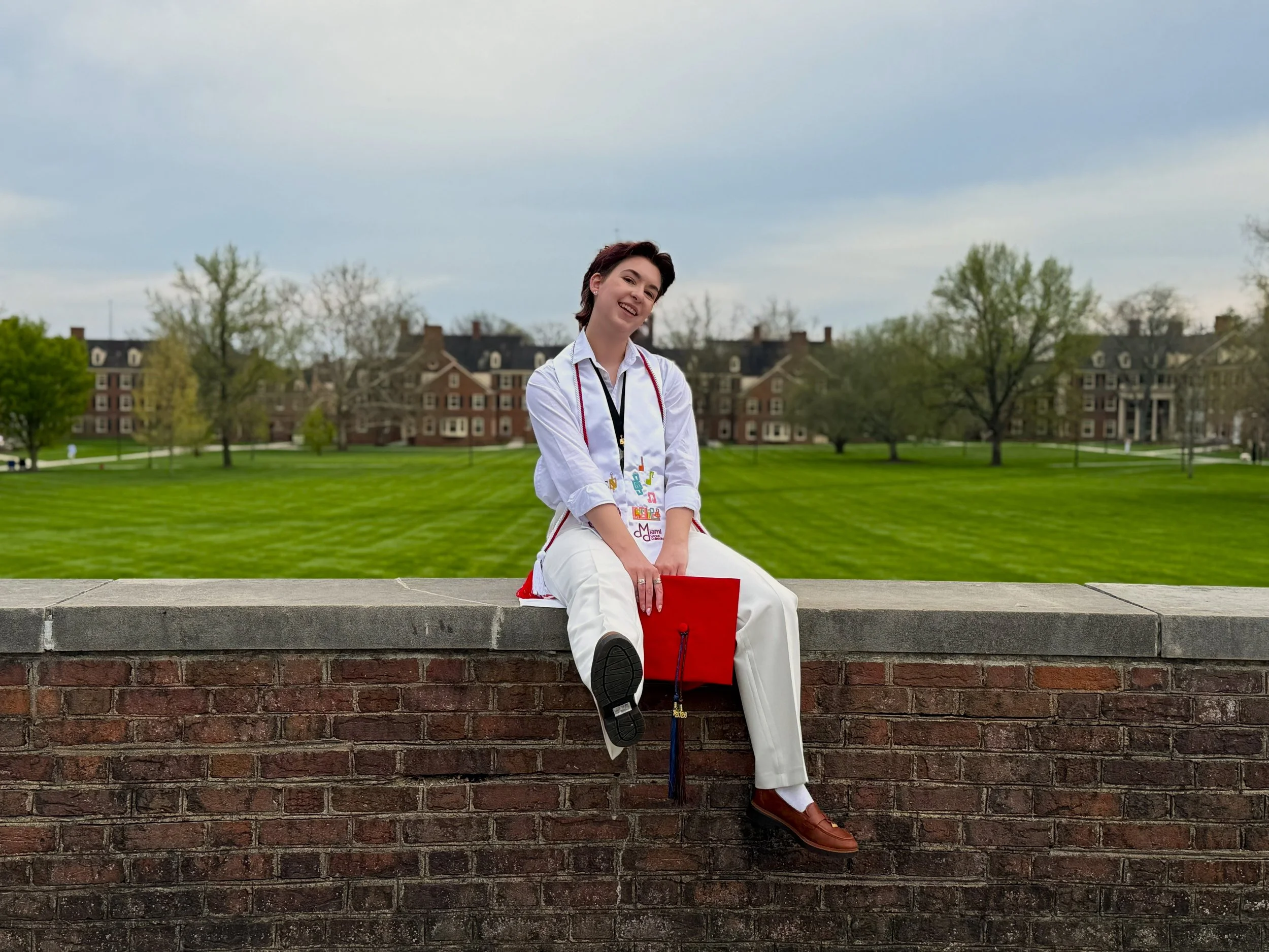 Founder, Courtney, sits on a brick wall outdoors on the Miami University campus, smiling and posing for a photo, with a grassy area and red brick buildings in the background.
