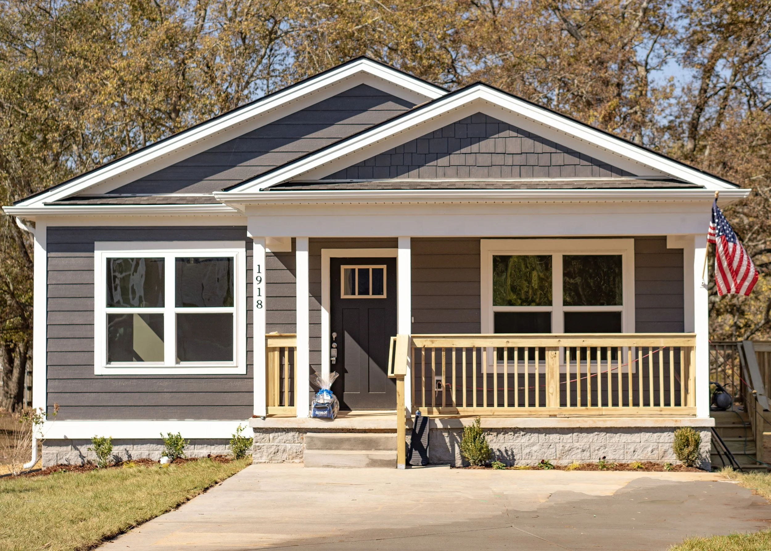 Single-story gray house for a housing advocacy organization, with a front porch, white trim, and an American flag, set against a backdrop of trees.