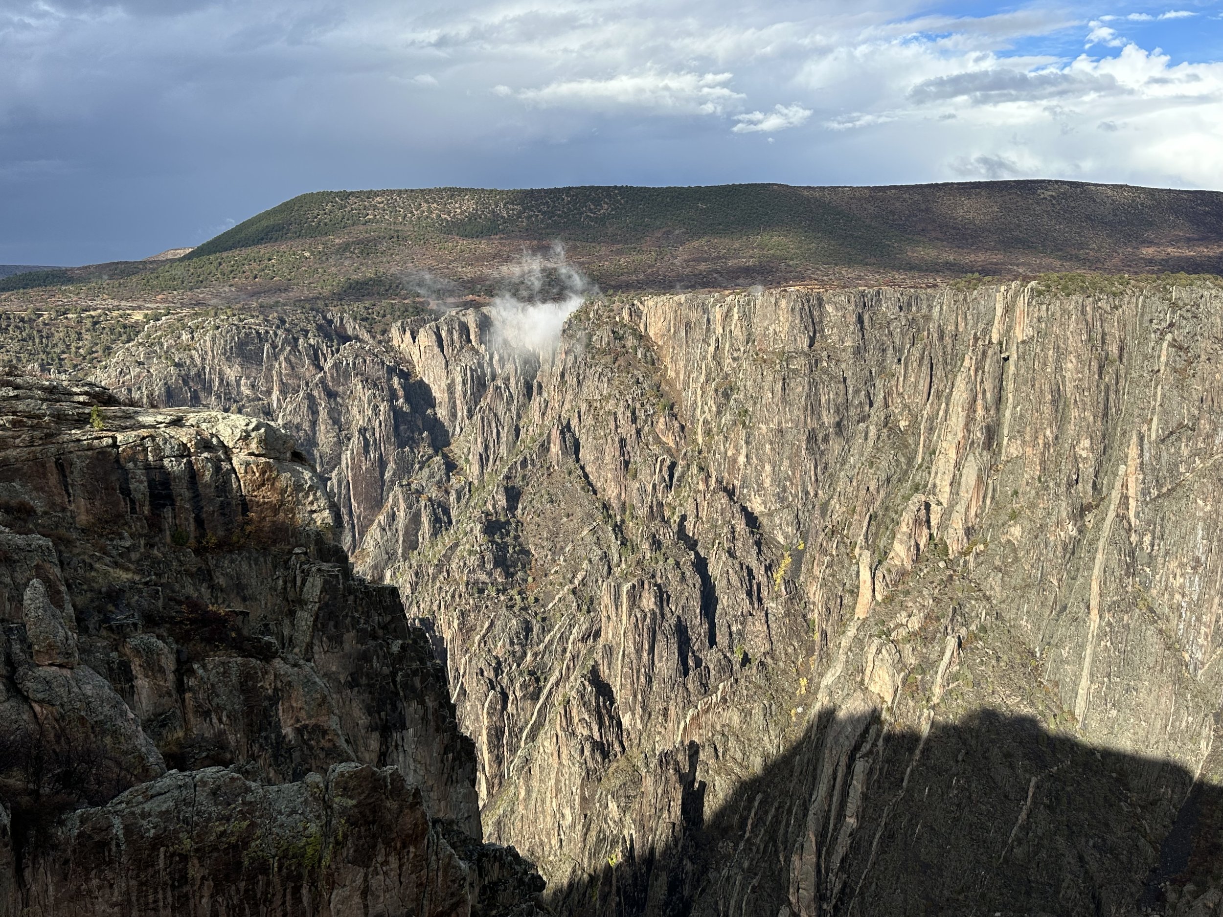 A deep canyon with steep rocky walls, steam rising from a hot spring or geyser, and a forested hill in the background under a cloudy sky.