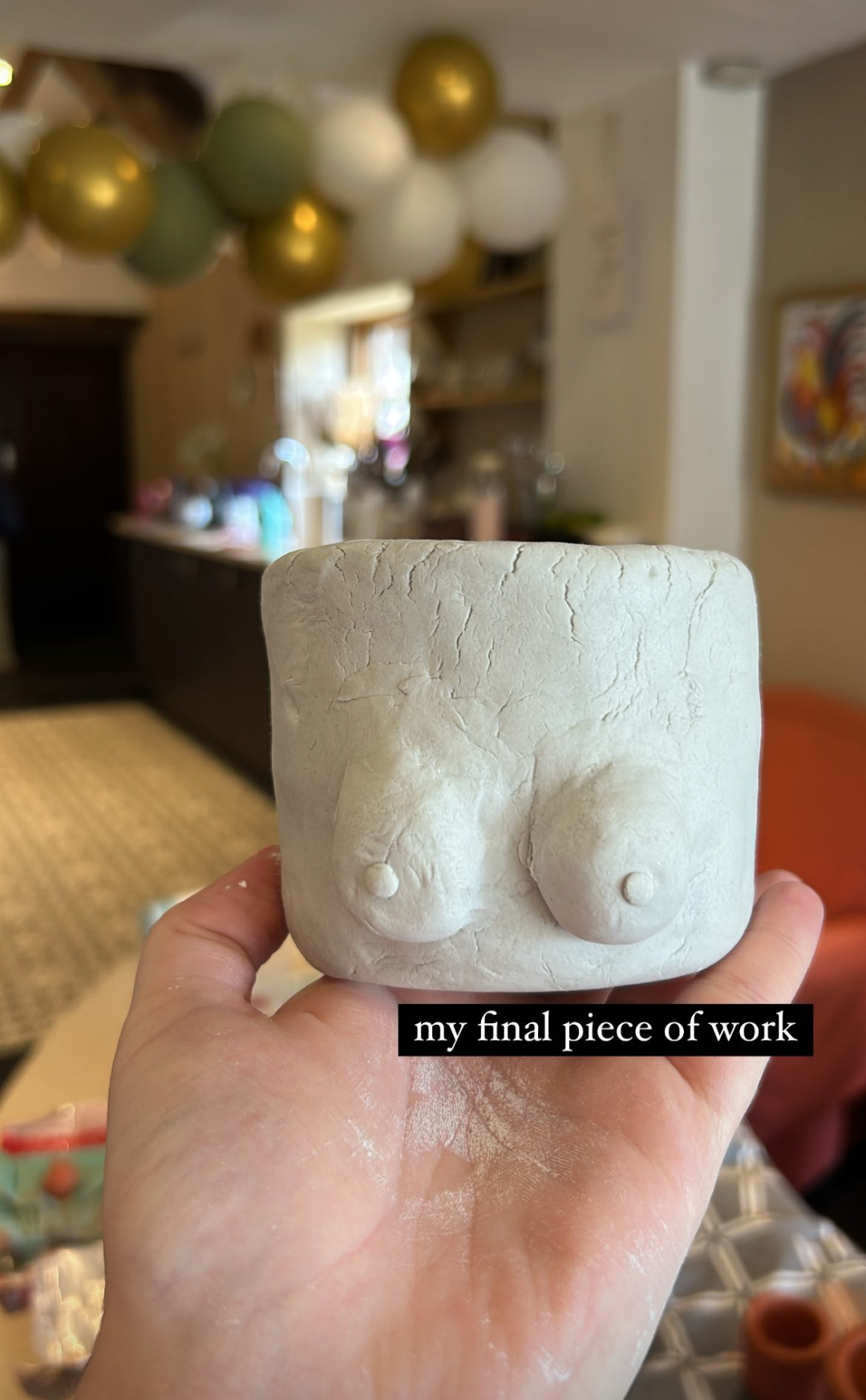 A person holding a small, unglazed ceramic sculpture of two breasts, with a kitchen and decorations in the background.
