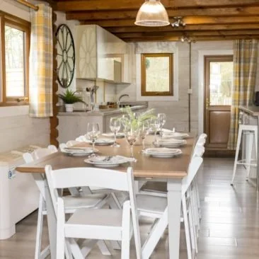Dining table set with plates, glasses, and a flower arrangement in a cozy, bright kitchen with rustic decor.