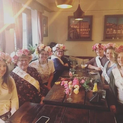 Group of women wearing flower crowns sitting around a table with flowers and drinks in a cozy, warmly lit room.