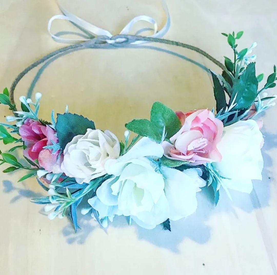 A floral headband with white, pink, and green artificial flowers and leaves, tied with a white ribbon.