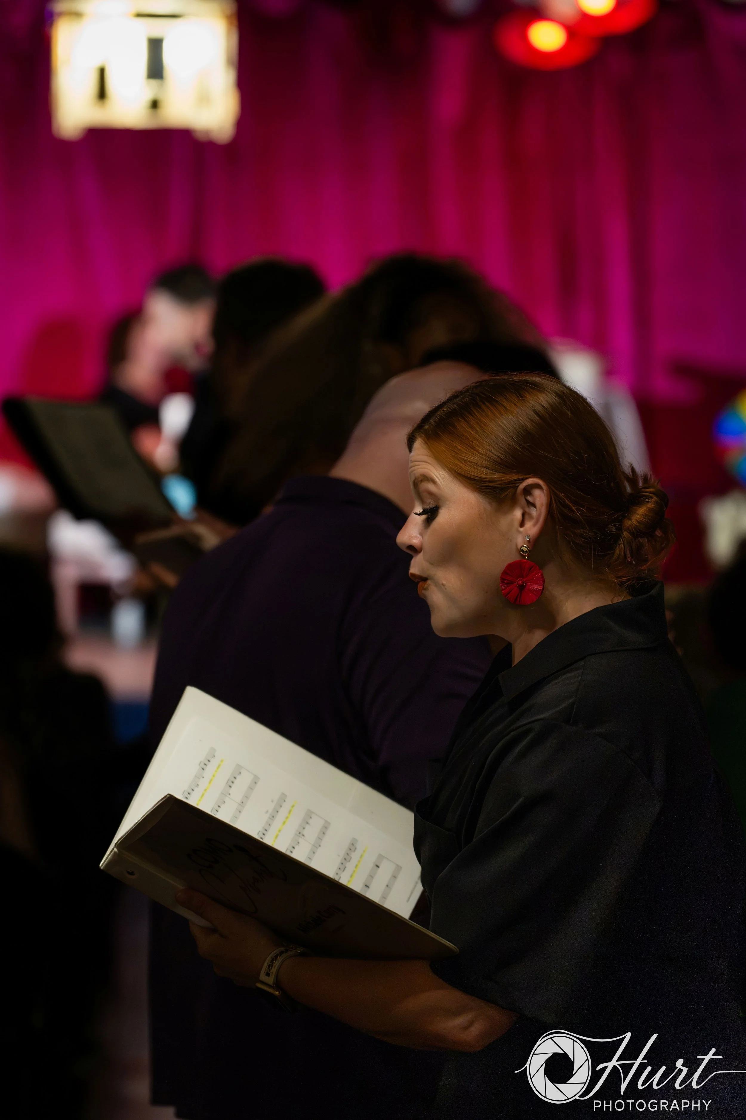 A woman with red hair and large red earrings is reading sheet music in a dimly lit venue with a purple background, other performers are visible in the background standing in a line.