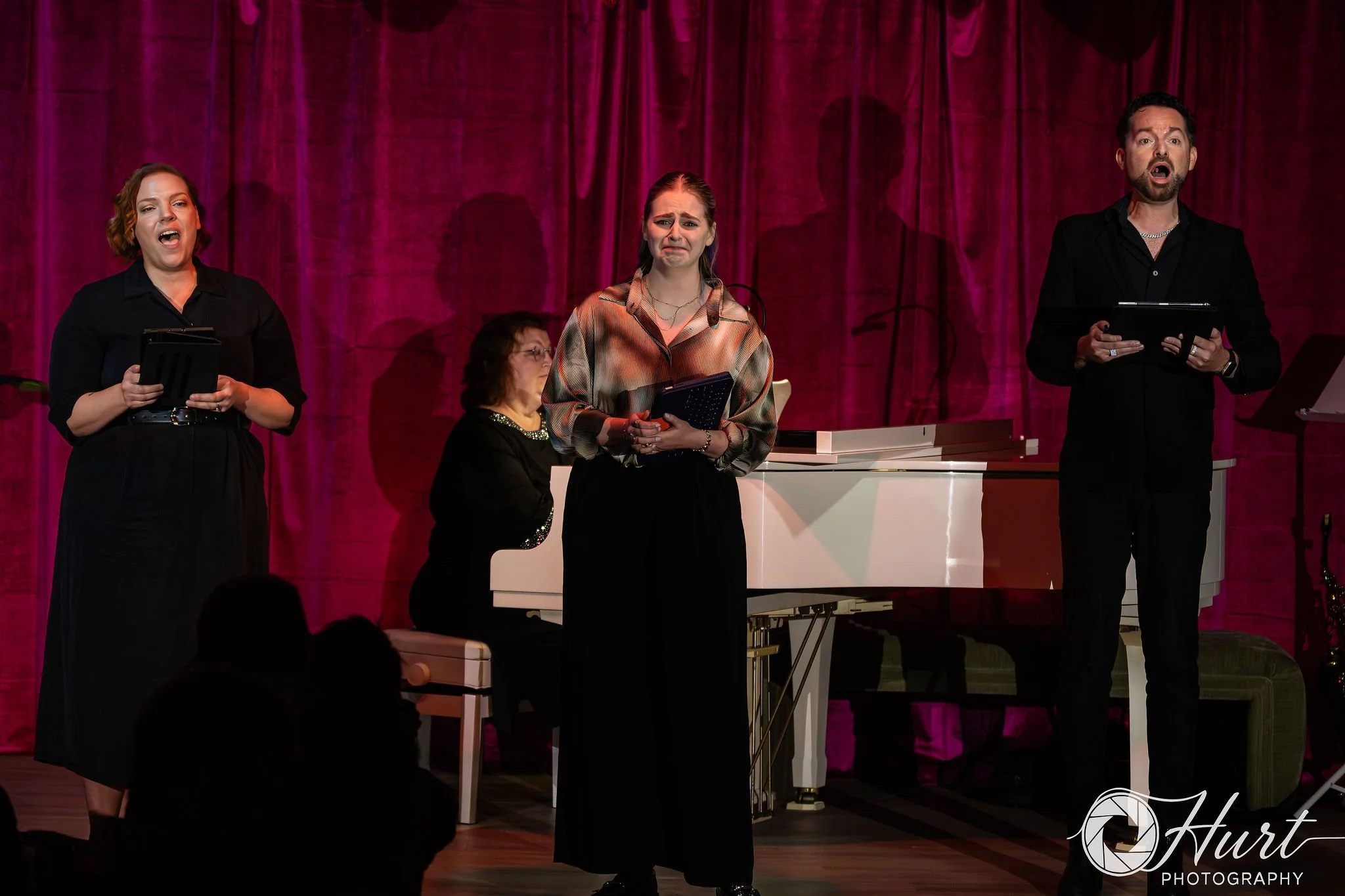 Three performers singing on stage, two women and one man, with a white piano and magenta curtain backdrop.