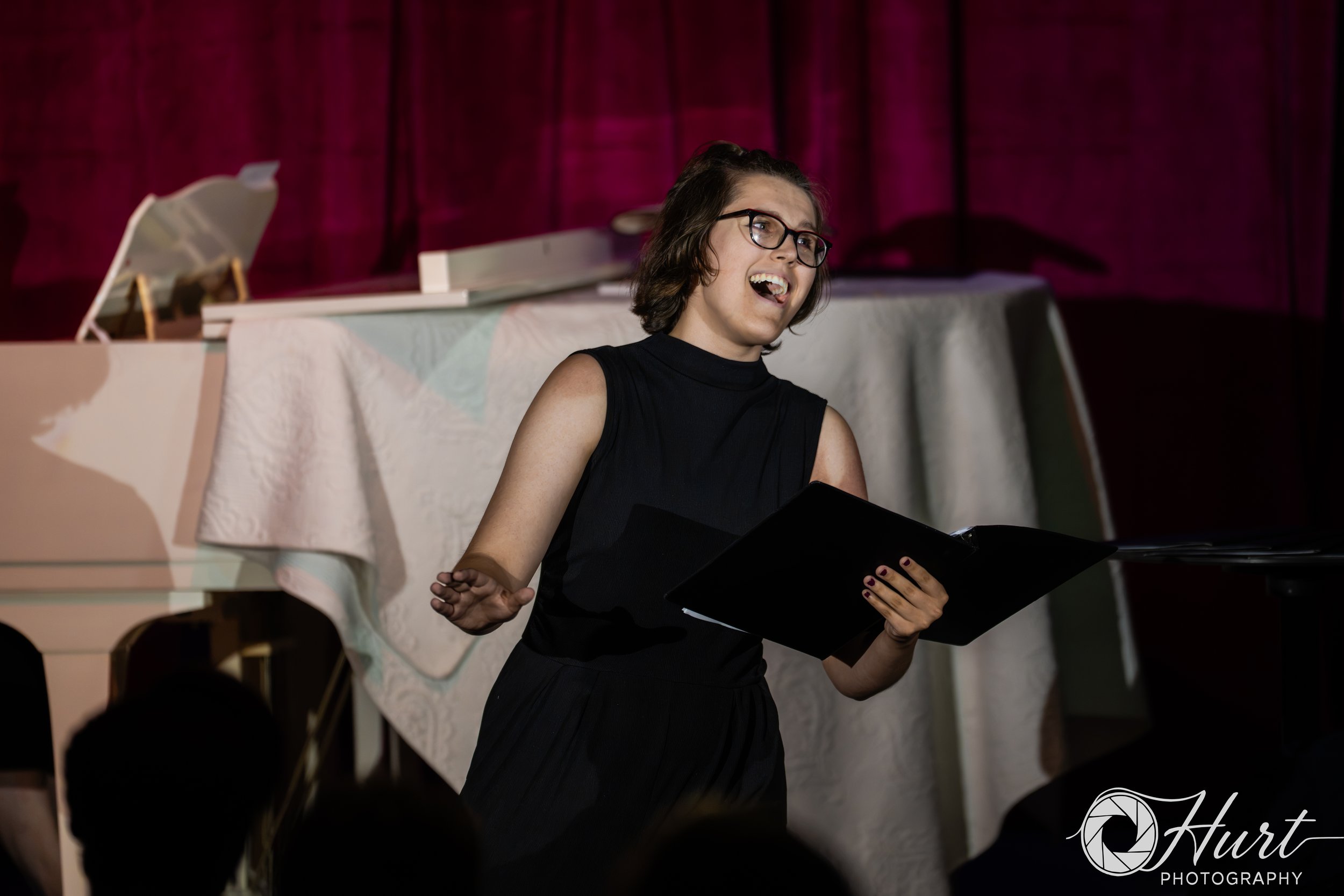 A woman with short brown hair, glasses, and a black sleeveless dress singing in front of a magenta curtain, holding a black folder or book in her left hand.