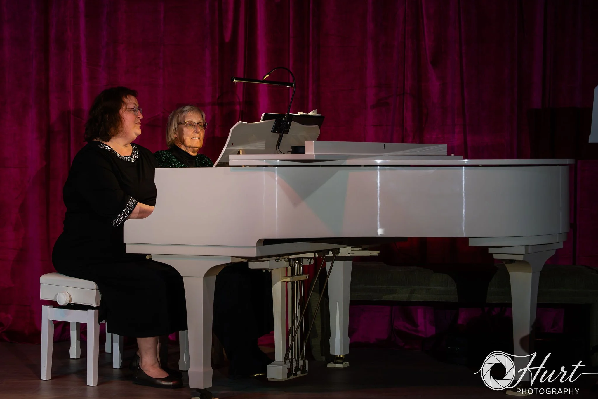 Two women sit at a white grand piano on a stage with a magenta curtain backdrop, both are playing the piano and dressed in black, with one woman wearing glasses.