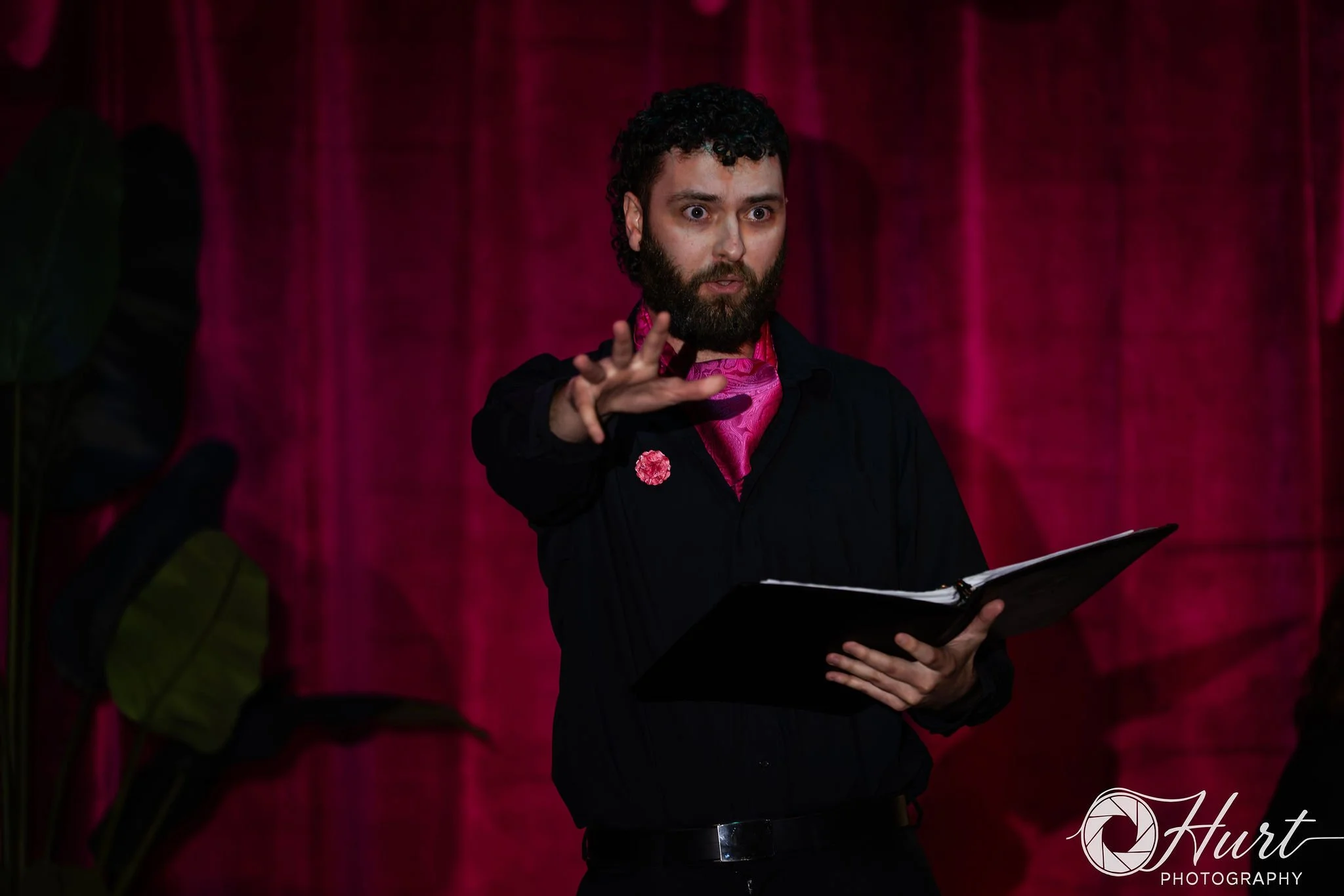 A man with a beard and curly hair wearing a black jacket and a magenta neckerchief, holding a tablet, standing in front of a magenta curtain, making a gesture with his right hand.