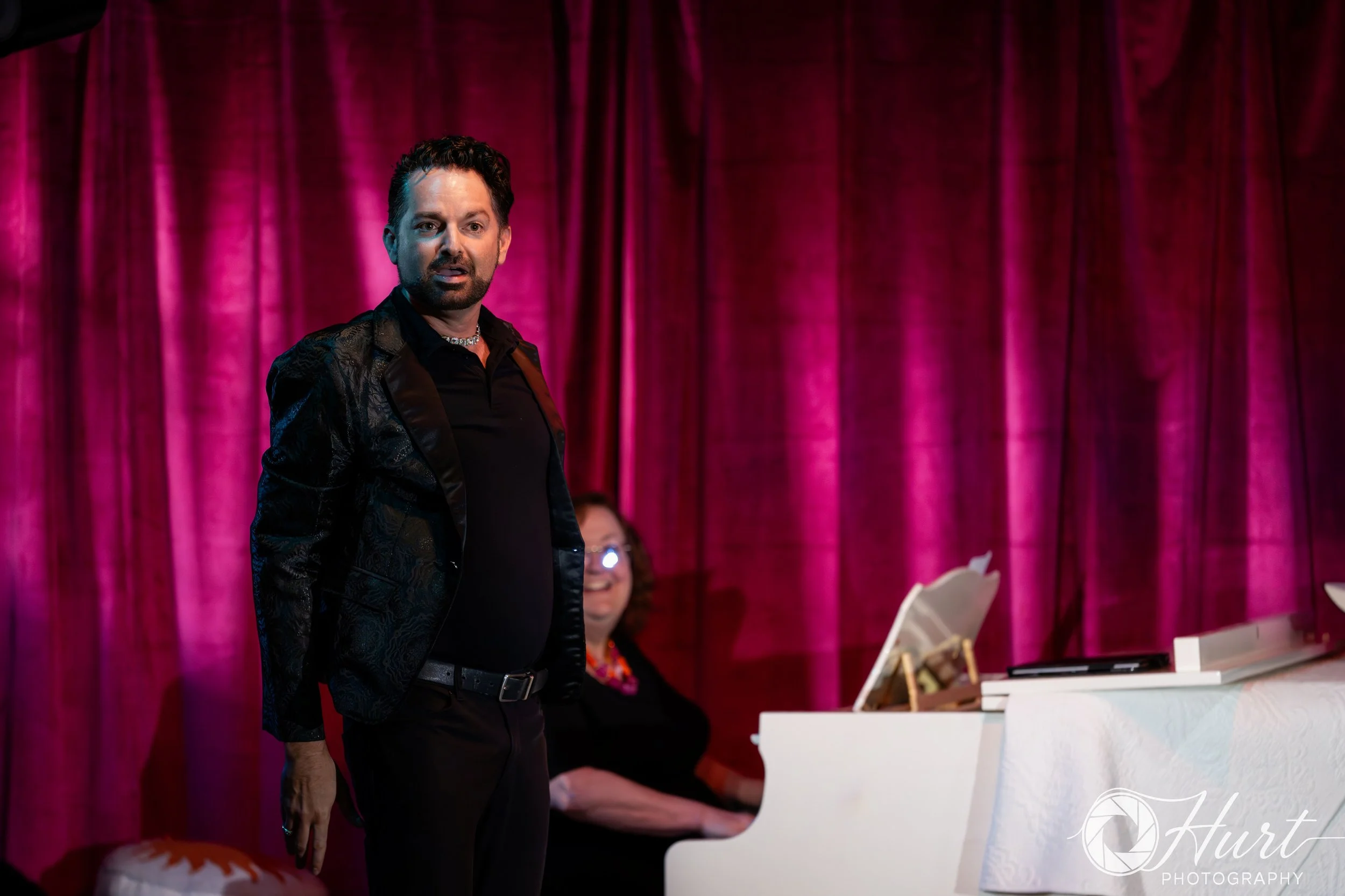 A man with dark hair, beard, and mustache wearing a black shirt and jacket stands singing in front of a magenta curtain, with a woman playing piano in the background.