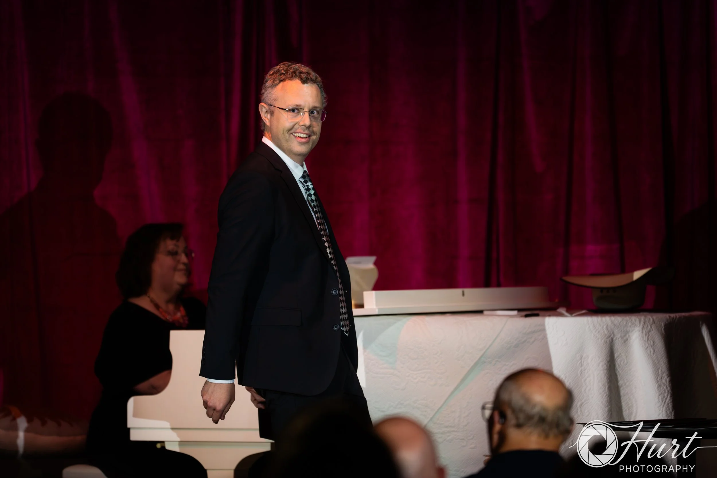 A man in a dark suit, collared shirt, and patterned tie dances on stage with a magenta curtain backdrop, smiling and looking at the camera, with people seated in front of him.